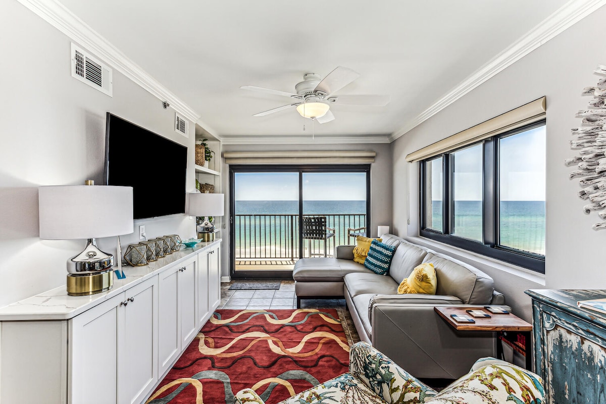 A stylish living area is highlighted by a large window offering expansive views of the Gulf. A grey sofa complements a vibrant area rug, while a mounted TV is positioned above a white cabinet. Natural light fills the space, enhancing the coastal ambiance.