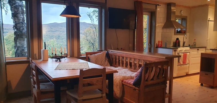 A dining area features a wooden table surrounded by chairs, complemented by a bench along the wall. Large windows provide expansive views of the surrounding landscape. The kitchen area is partially visible, showcasing modern appliances and wood cabinetry, enhancing the bright and open feel of the space.