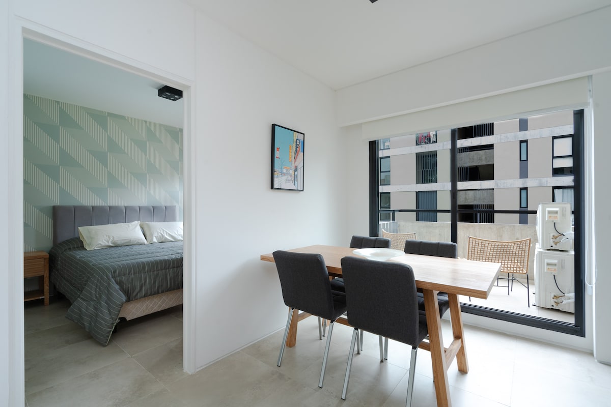 A bright living and dining area showcases a wooden dining table with four chairs. A spacious bedroom is visible through an open doorway, featuring a large bed with a neatly arranged quilt. Natural light filters in through large windows, illuminating the modern design.