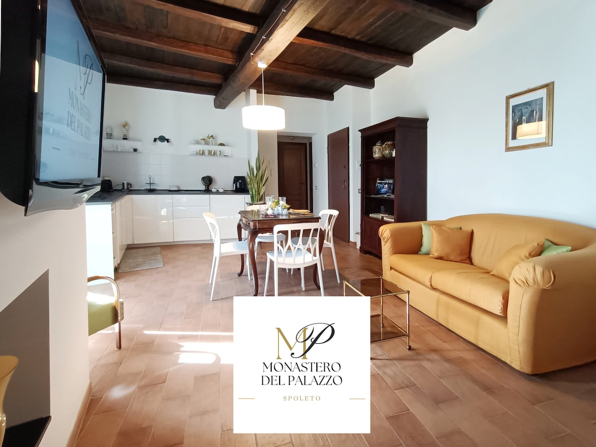 A bright living area features a comfortable yellow sofa and a dining table surrounded by white chairs. The modern kitchen is visible in the background, featuring sleek cabinetry and essential appliances. Natural light floods the space, highlighting the wooden ceiling beams and warm tiled flooring.