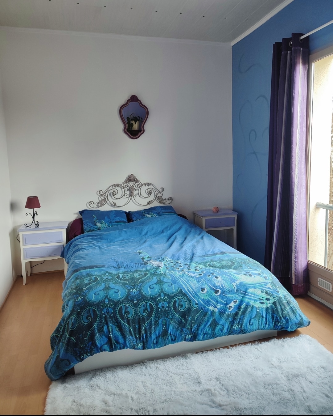 A spacious bedroom features a bed dressed in vibrant blue bedding with peacock designs. Nightstands are positioned on either side of the bed, complementing the room's decor. A large window is partially covered by dark curtains, allowing soft light to filter in.