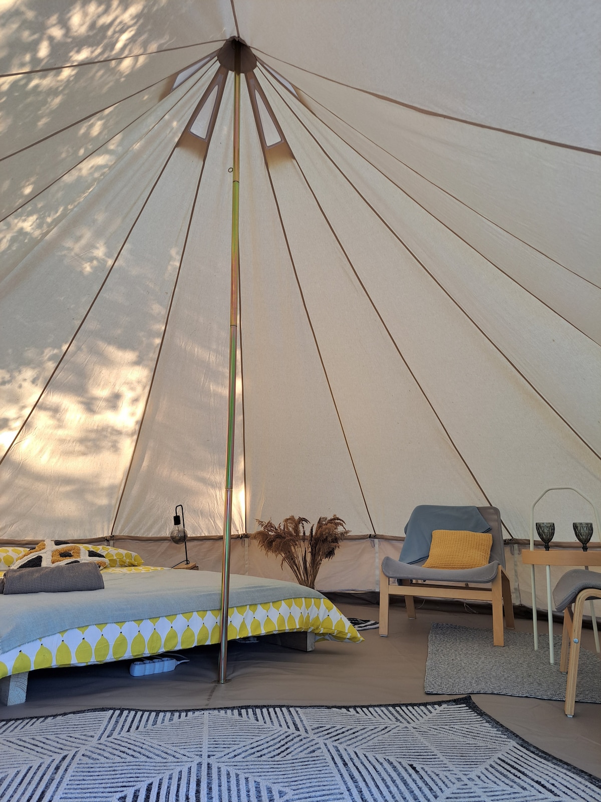 A spacious glamping tent features a large bed adorned with a patterned duvet and cozy towels. Simple seating includes a wooden chair and a rug on the ground. Natural light filters through the tent fabric, creating a calming atmosphere.