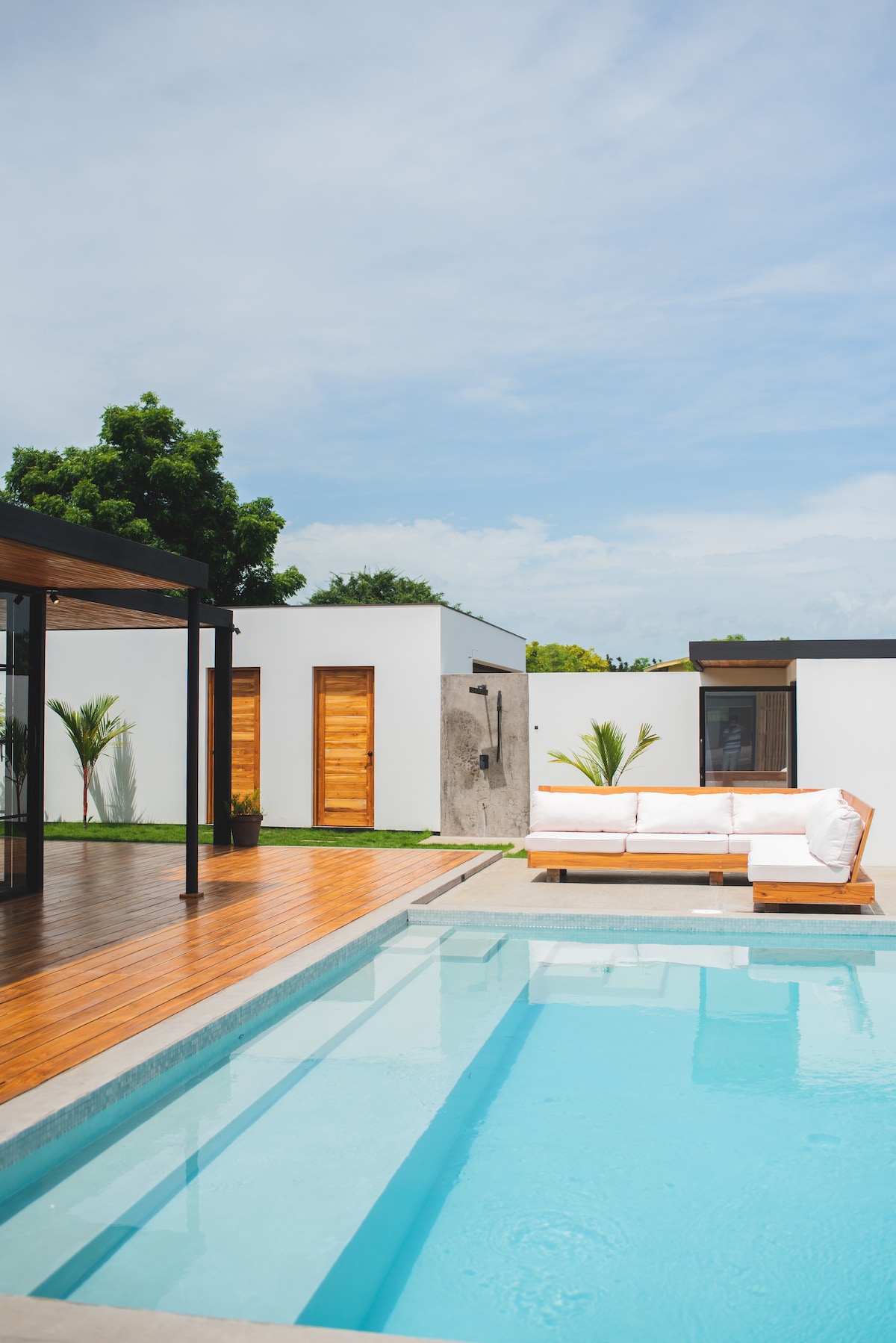 A clean, minimalist pool area features a light-colored sectional sofa positioned on a wooden deck next to a refreshing turquoise pool. Nearby, white walls and wooden doors create a serene backdrop, while green plants add a touch of nature to the space.