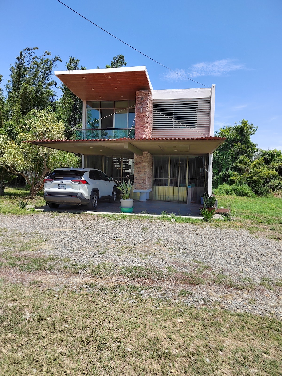The modern two-story house features a combination of stone and glass, surrounded by greenery. A driveway is visible in front, accommodating a parked vehicle. The upper level is accented by a wooden overhang, providing shade, while large windows invite natural light inside.