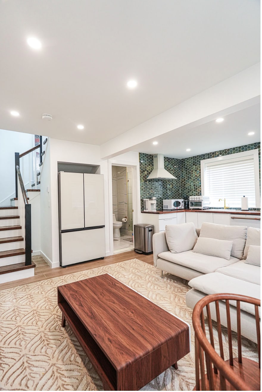 A bright living area is presented, featuring a light-colored sofa and a wooden coffee table. A staircase leads to the upper level, while a modern kitchen with a distinctive tiled backsplash is visible in the background. Natural light illuminates the space through large windows.