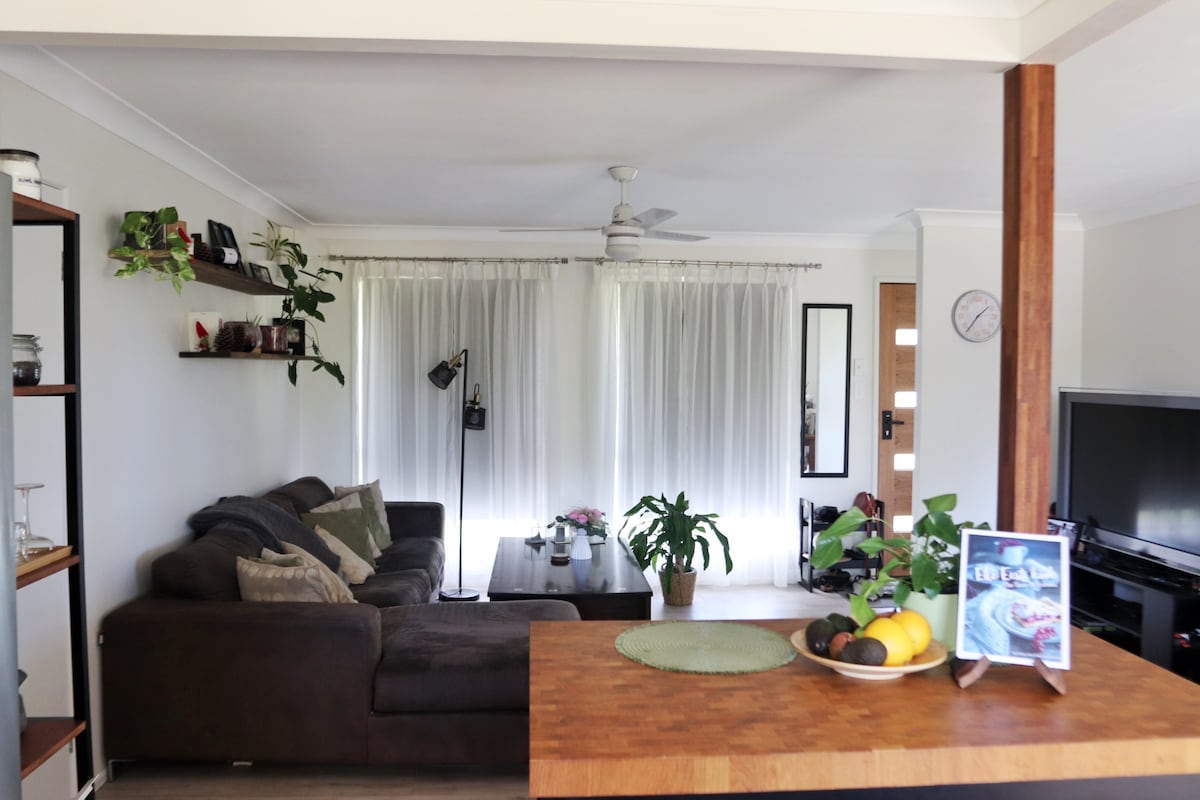 An inviting living area features a comfortable dark sofa adorned with cushions. A coffee table is set with a bowl of fruit, while plants add freshness. Sheer curtains frame the windows, allowing soft light to illuminate the space, complemented by a floor lamp and a mounted clock.