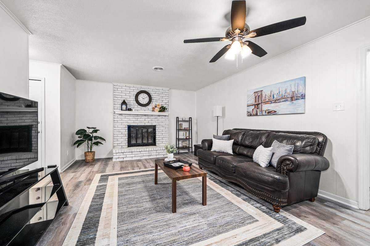 A cozy living space features a leather sofa positioned across from a modern television. A brick fireplace serves as a focal point, complemented by a decorative clock above it. A textured area rug covers the floor, adding warmth, while a ceiling fan provides airflow.