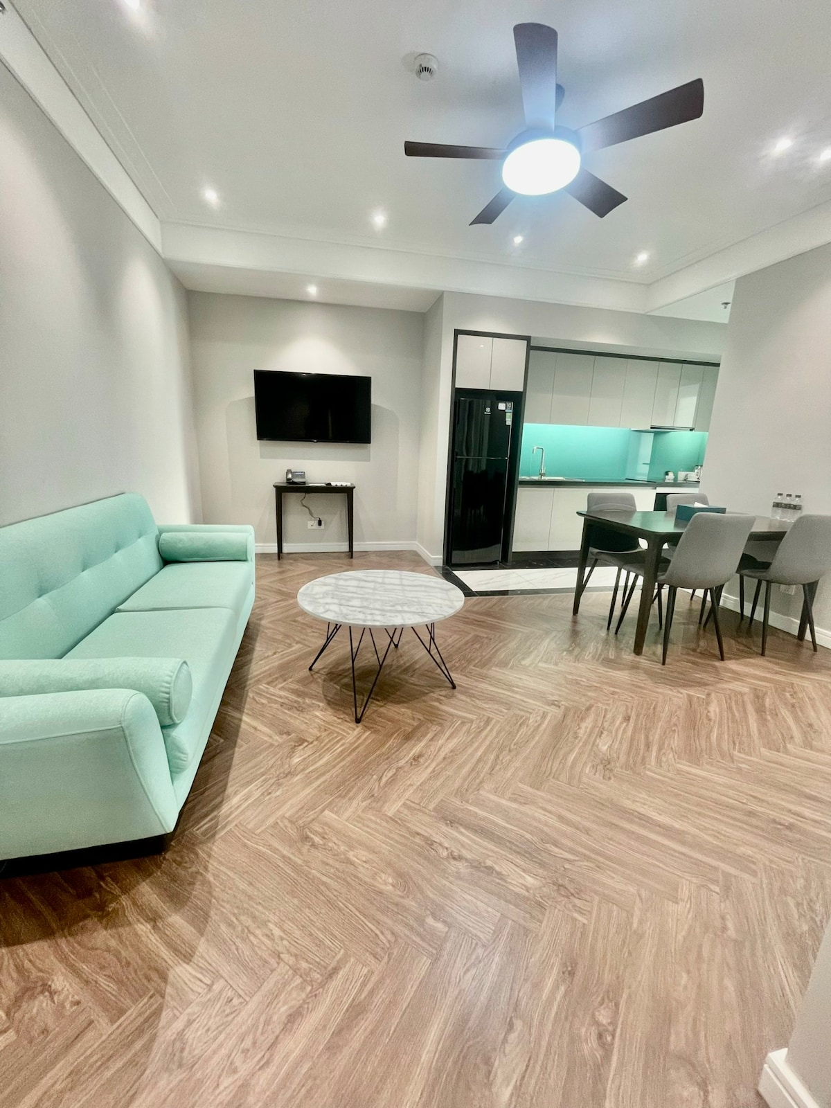 A modern living area features a light green sofa positioned in front of a flat-screen television. A round coffee table sits on a wood-patterned floor, accompanied by a dining set in the background. The kitchen area, outlined in teal, showcases contemporary appliances.