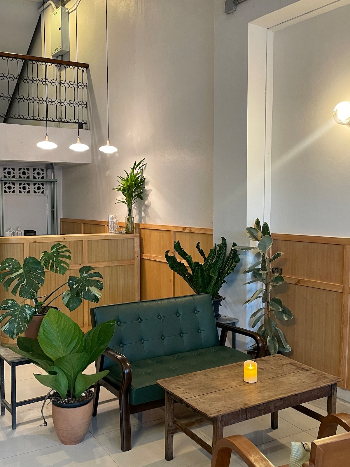 A welcoming seating area is present with a green upholstered sofa and a rustic wooden table. Several potted plants add greenery to the space. Soft lighting is provided by pendant fixtures overhead, contributing to a calm ambiance.