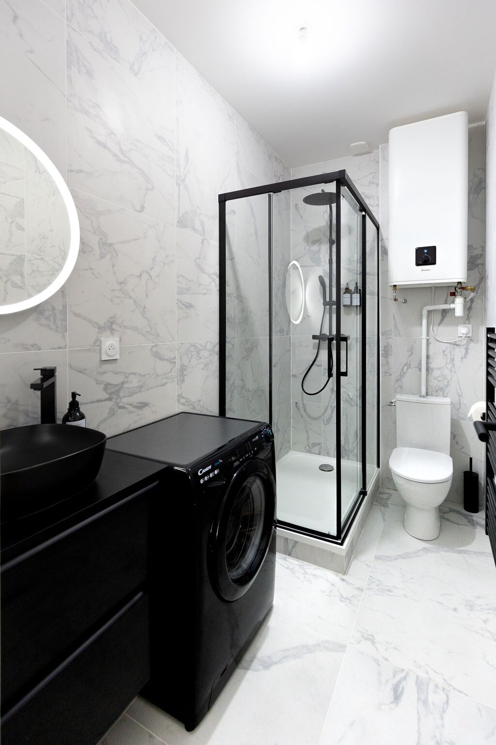 A modern bathroom is equipped with a glass shower enclosure, a sleek black basin, and a washing machine. Light marble tiles cover the wall and floor, while a circular mirror adds a touch of elegance. A white toilet is situated beside the shower.