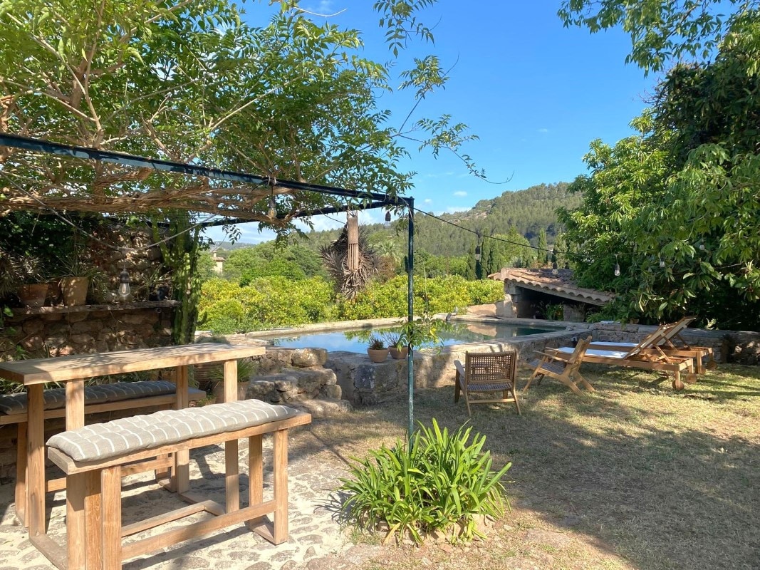 An outdoor relaxation area is featured, with a wooden table and benches accompanied by lounge chairs. A verdant landscape surrounds a natural well, providing a serene backdrop. Trees and bushes offer shade while the clear sky enhances the tranquil atmosphere of the space.