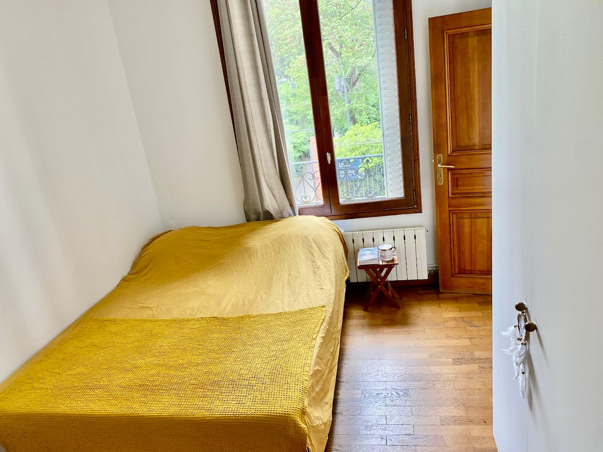 A private bedroom features a spacious bed covered with a bright yellow blanket. Natural light filters through large windows framed by light curtains, revealing a glimpse of greenery outside. A small wooden table is positioned beside the bed, enhancing the room's functionality.