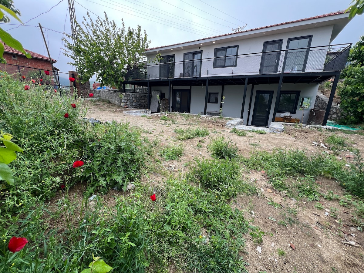 A modern two-story stone house is surrounded by a garden featuring green grass and blooming wildflowers. The structure has a deck supported by black pillars, providing elevation. The exterior showcases large windows and a blend of light and dark colors, creating a harmonious visual appeal.