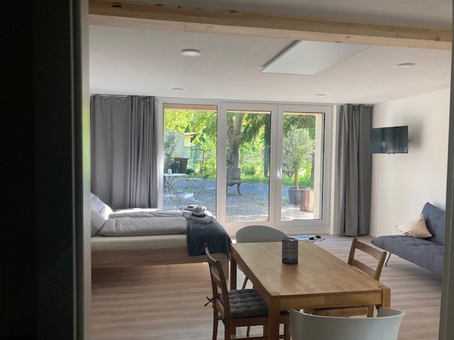 A spacious living area features large glass doors that open to a green outdoor space. A comfortable bed is positioned near the window, while a dining table and chairs are arranged thoughtfully across the room. A seating area is visible in the corner.