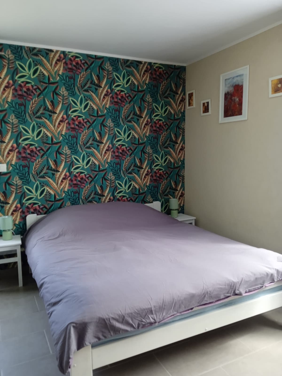 A cozy sleeping area features a spacious bed covered with a lavender sheet. Colorful botanical wallpaper adorns one wall, complemented by two bedside tables with lamps. Small framed artworks add a touch of decor, enhancing the room's inviting ambiance.