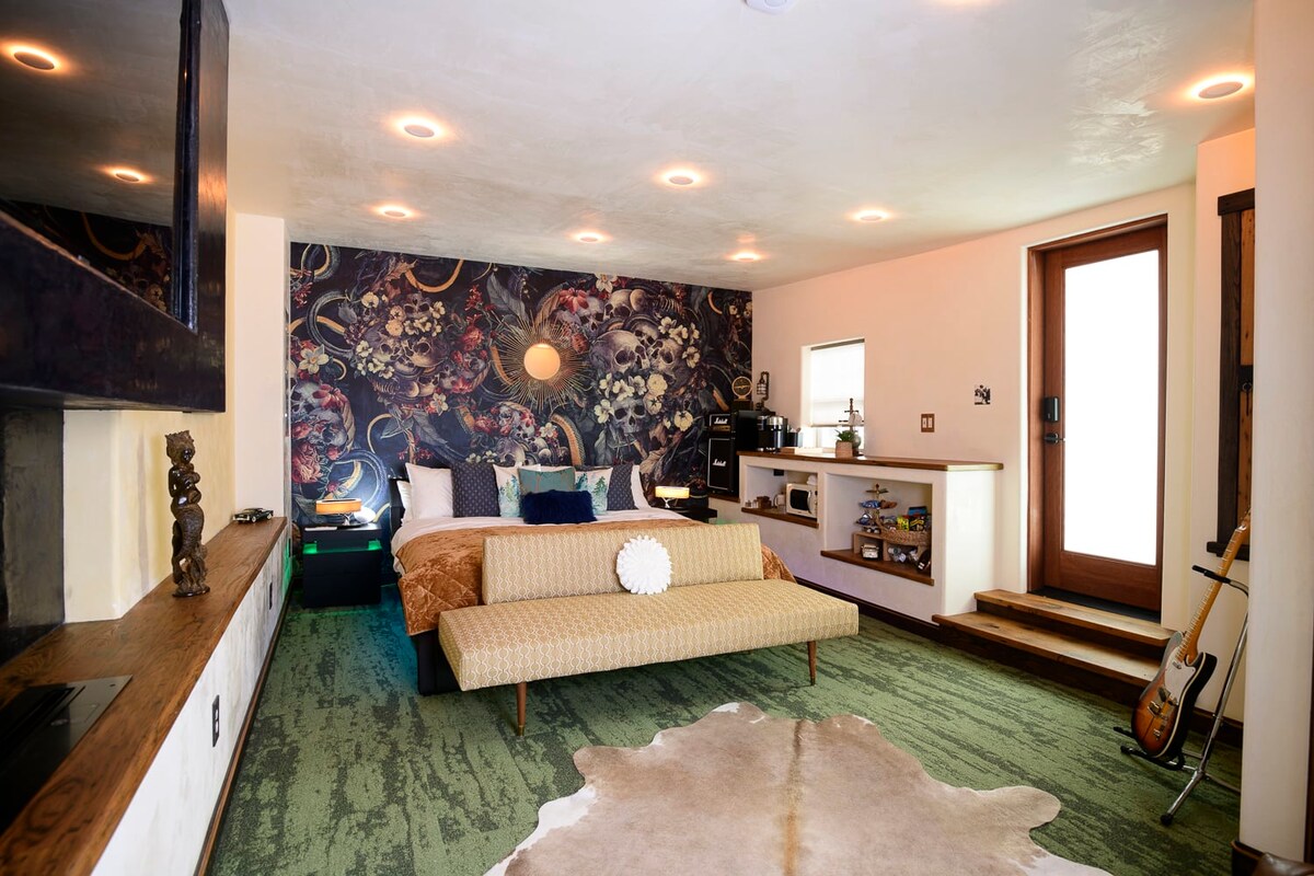 A spacious bedroom is presented, featuring a large bed with patterned bedding. A decorative bench is positioned at the foot of the bed. Artistic wall decor adds character, while natural light enters through a nearby door. A cozy area rug complements the overall aesthetic.