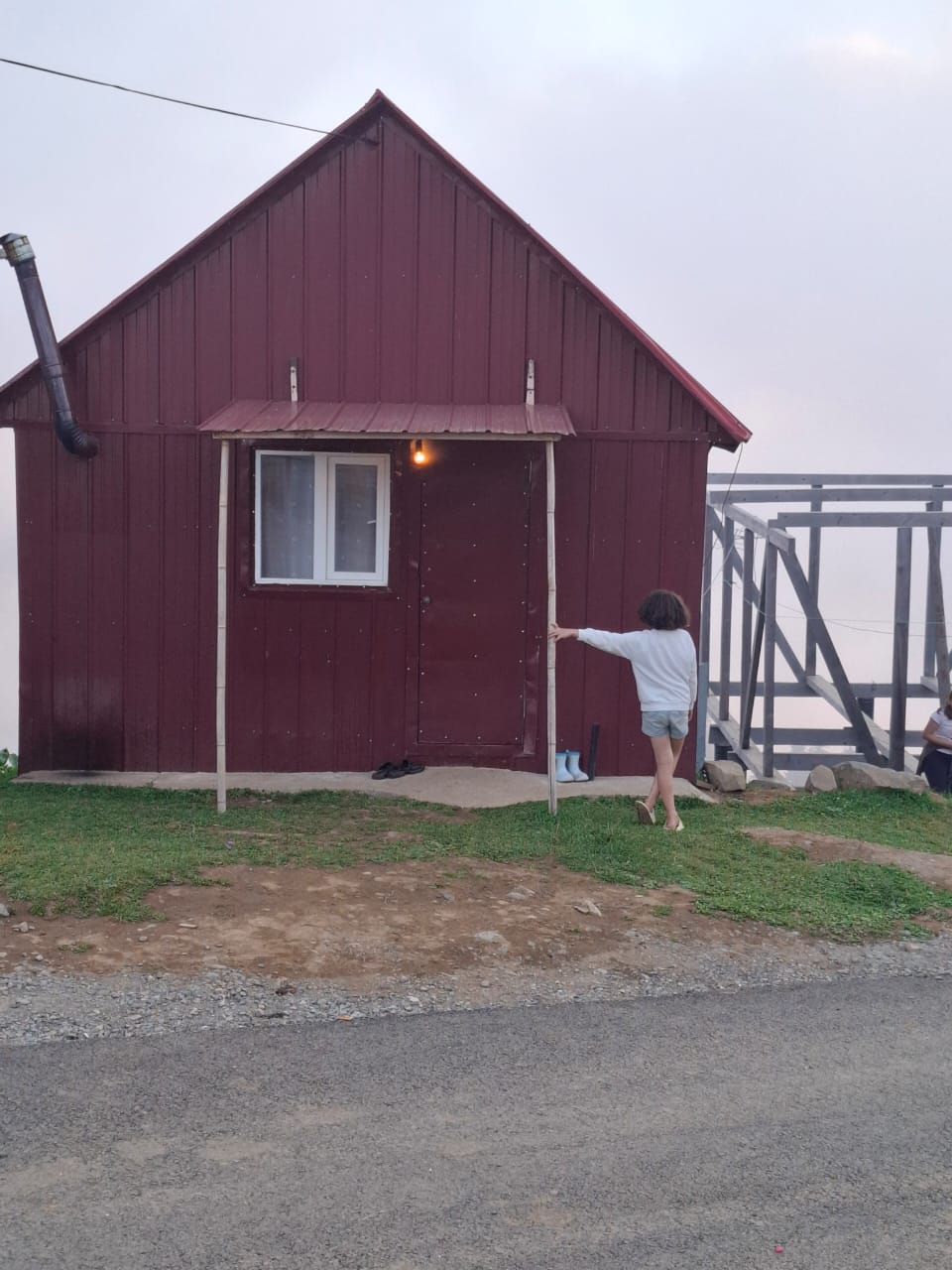 A small, red cabin with a gabled roof is positioned near a gravel road. A single window allows light to filter in, while a soft glow emanates from the door. A person is seen approaching the entrance, surrounded by grassy terrain.