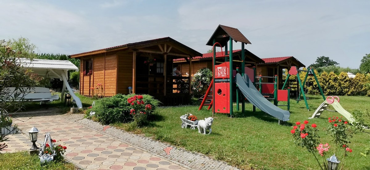 A wooden cabin structure appears alongside a diverse outdoor play area, featuring a slide and swings designed for children. A well-maintained garden with colorful flowers, a stone pathway, and decorative elements completes the inviting exterior, well-suited for families seeking outdoor activities.