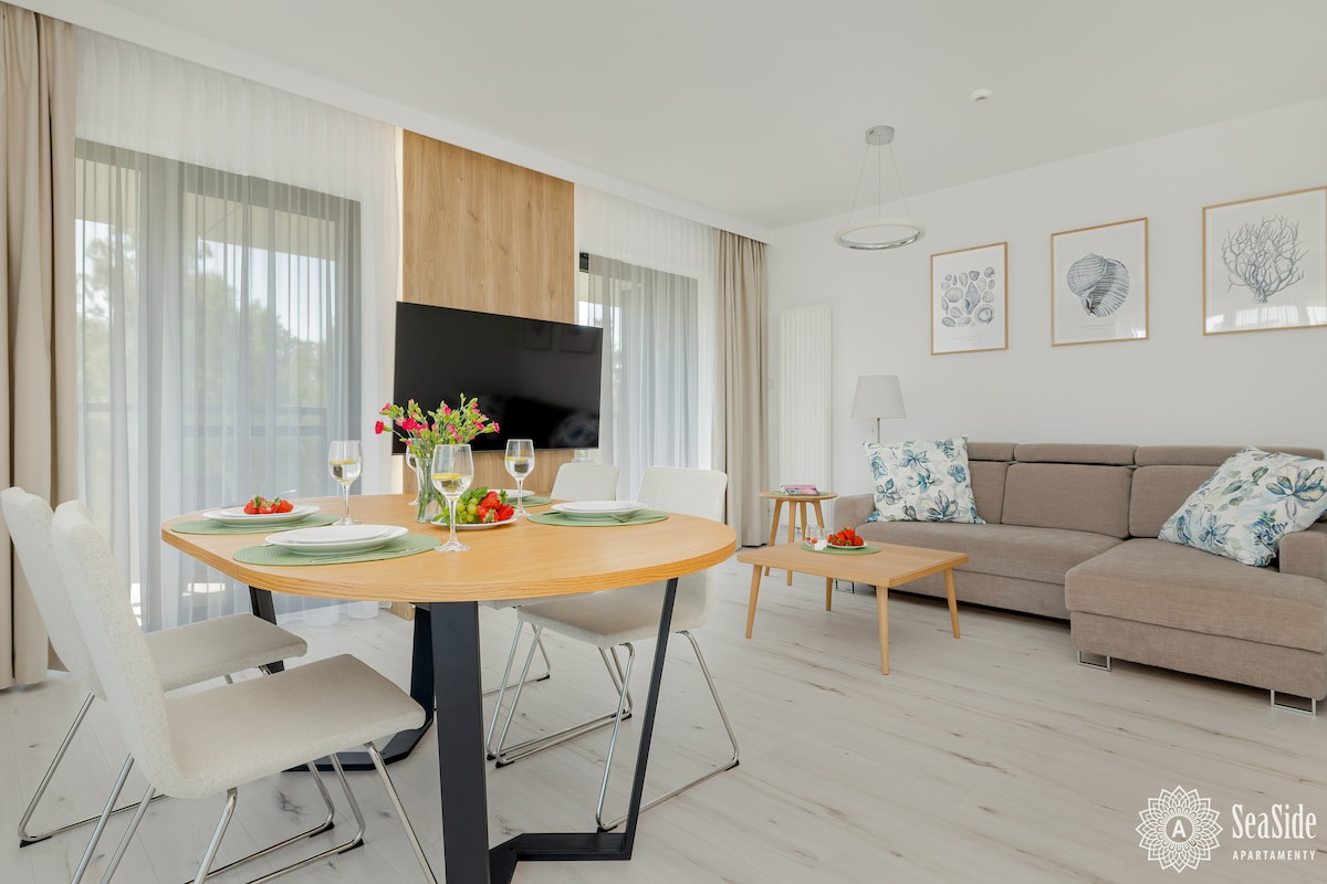 A modern living area is featured, showcasing a round dining table set for two with stylish chairs. A comfortable sofa sits nearby, complemented by a side table. Large windows allow natural light to fill the space, with soft curtains enhancing the ambiance.