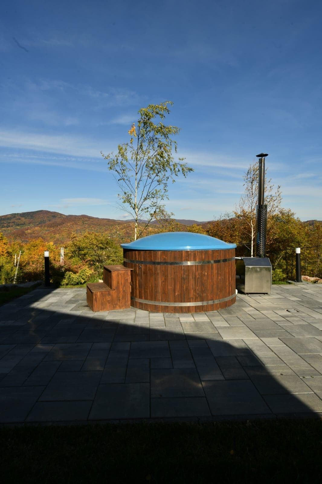 A wooden hot tub is positioned on a stone patio, surrounded by autumn foliage and mountain views. A small stone chimney is adjacent to the hot tub, contributing to a serene outdoor atmosphere. Soft sunlight bathes the scene, highlighting the natural landscape.