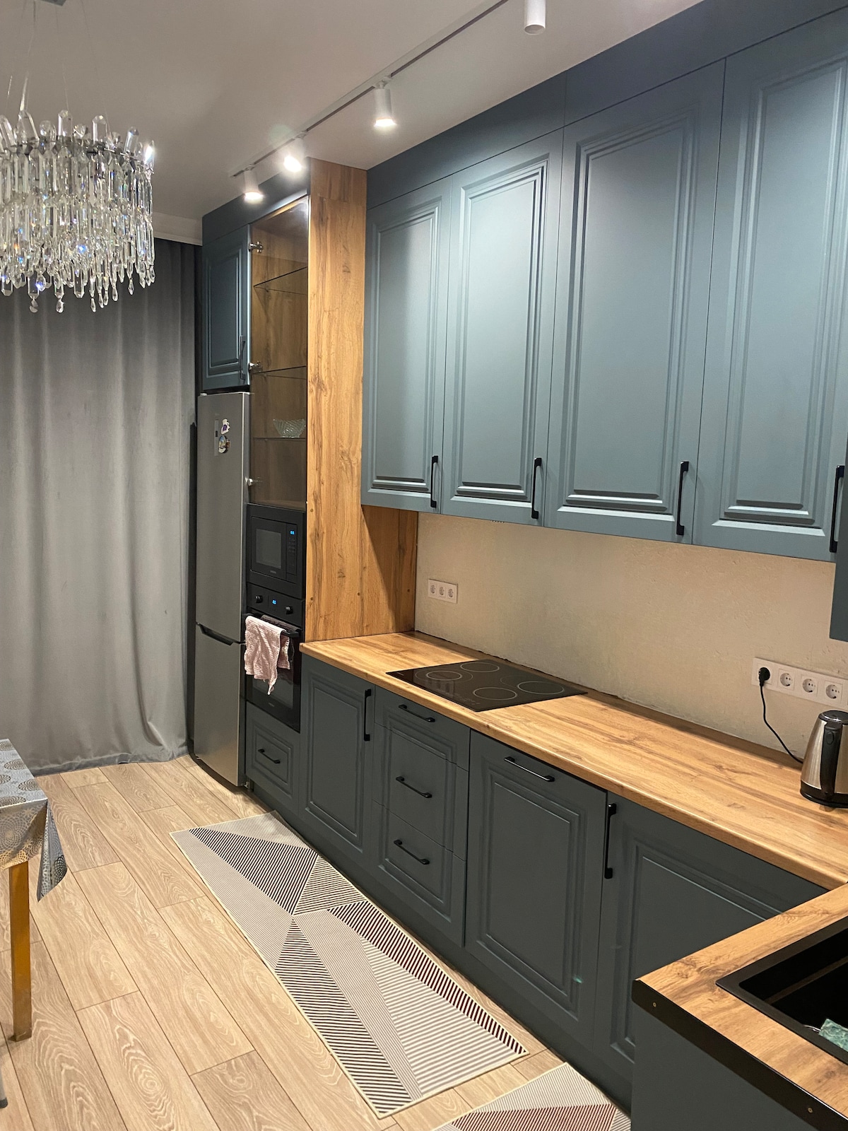 A modern kitchen featuring dark blue cabinets with a wooden accent wall. Stainless steel appliances, including a refrigerator and microwave, are visible. A glass chandelier hangs above, providing additional lighting. The countertop is spacious, and a patterned rug lies underfoot.