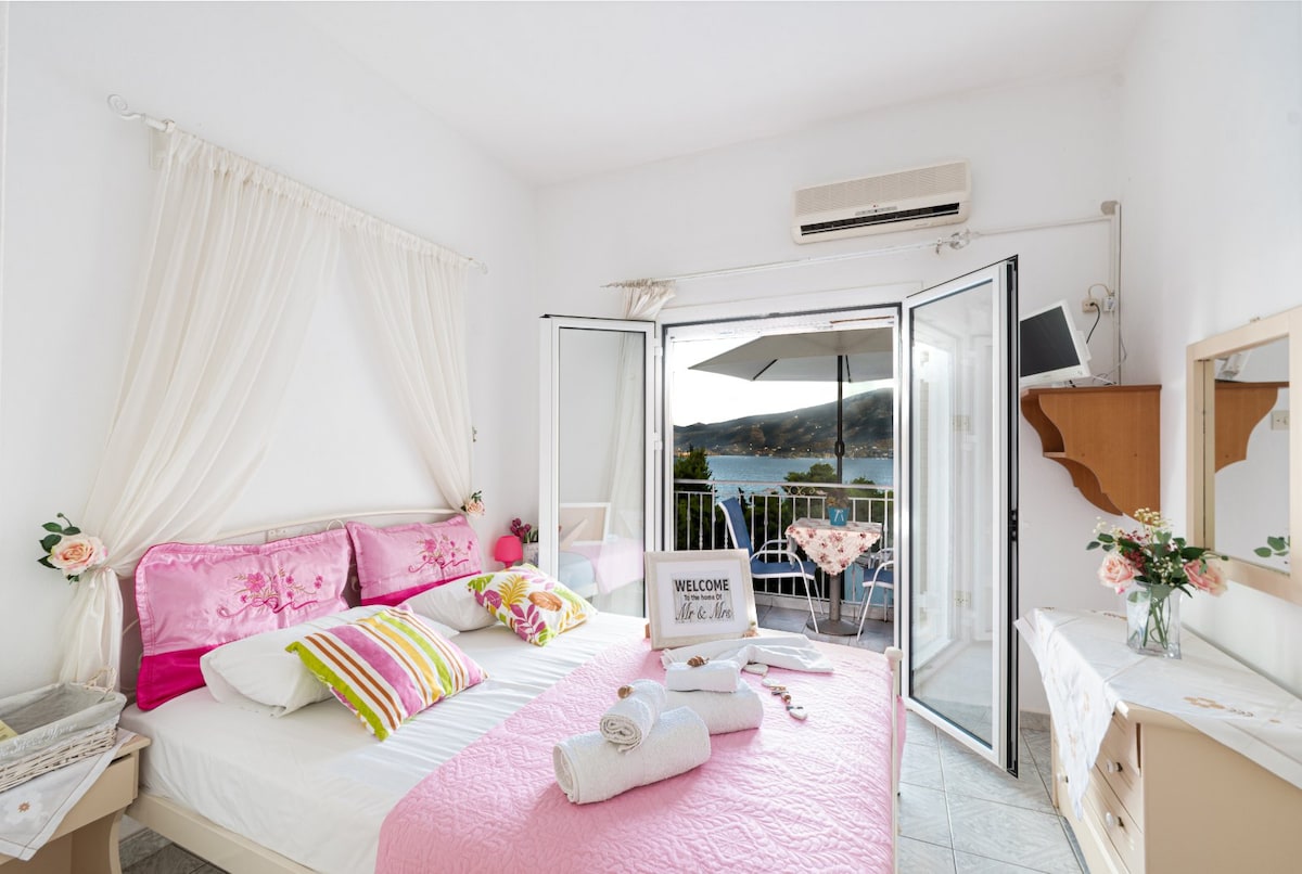 A bright bedroom features a spacious bed adorned with pink and white textiles. Natural light floods in through double doors, leading to a balcony with views of the water. A vanity and dresser provide additional storage, complemented by decorative flowers.