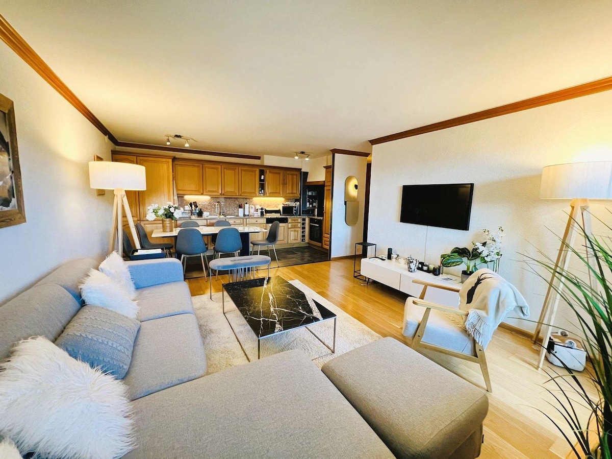 A spacious living area features a light-colored sectional sofa and a sleek black coffee table, with a dining space set for four. The kitchen is visible in the background, showcasing wooden cabinetry and modern appliances. Warm lighting creates a comfortable environment.