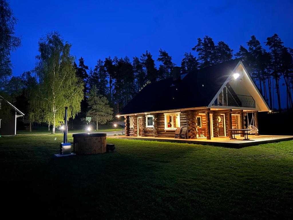 A log house is illuminated at night, revealing a charming façade and inviting outdoor seating. Soft lighting highlights the surrounding greenery, including a tree and grass. A wooden hot tub is visible on the patio, providing a serene outdoor space.
