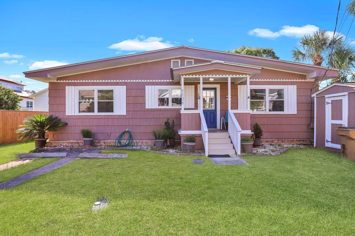 The exterior of the cottage is showcased, featuring a welcoming porch with white railings and a blue front door. Light pink siding complements the white shutters on the windows. A lawn with green grass and landscaping elements surrounds the cottage, creating an inviting atmosphere.