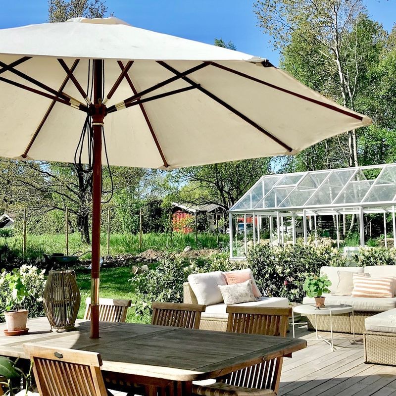 A spacious wooden deck is sheltered by a large umbrella, providing shade for a dining table surrounded by chairs. In the background, a greenhouse is visible alongside lush greenery, enhancing the outdoor setting. Potted plants add natural beauty to the tranquil space.