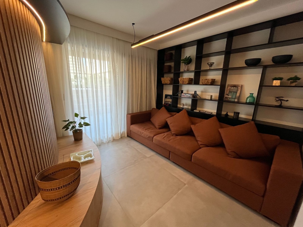A cozy sitting area features a spacious brown sofa with a variety of decorative pillows. Shelving units with plants and decorative items are arranged against the wall, while soft natural light filters through sheer curtains, providing a calm ambiance.