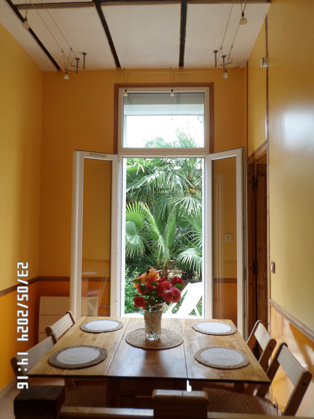 A dining area is filled with natural light from large windows that open to a private garden. A wooden table is set with placemats and a vase of vibrant flowers, creating a fresh and inviting atmosphere. The walls are painted in a warm yellow tone.