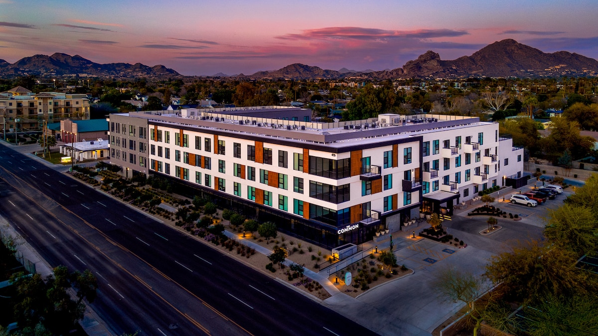 A modern apartment complex is depicted, featuring a sleek exterior design with a mix of light and dark elements. Surrounding mountains provide a natural backdrop, while the roadway and landscaped areas add to the overall setting. The image captures a tranquil evening sky with soft pastel colors.