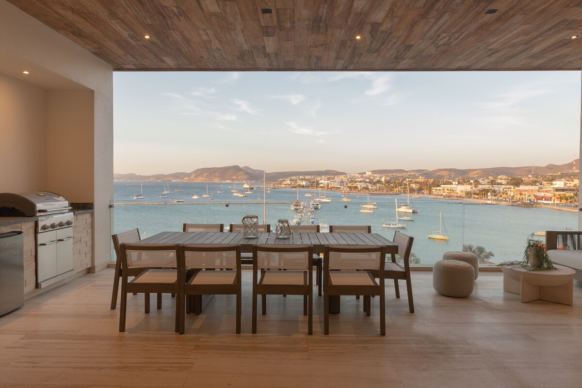 An outdoor dining area is presented, featuring a spacious table surrounded by several chairs. A BBQ grill is visible adjacent to the dining setup. The expansive glass wall reveals stunning views of the ocean and boats, enhancing the setting's coastal appeal.