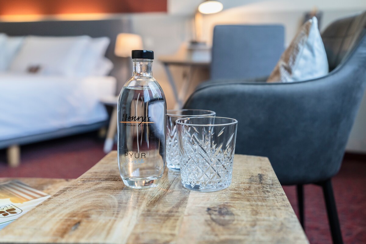 A bottle of water sits on a wooden table alongside two crystal-clear glasses. In the background, a spacious bed is neatly made with white linens, and a lamp provides soft lighting in the cozy setting.
