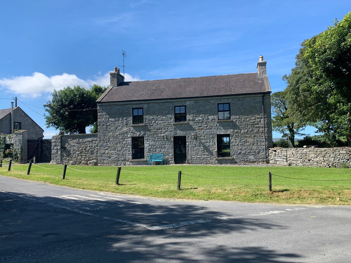 The historic country house of stone is framed by lush greenery, with a well-maintained lawn and a clear blue sky overhead. Large windows reflect the surrounding environment, and a patio area is visible in the front, enhancing the inviting exterior of this spacious dwelling.