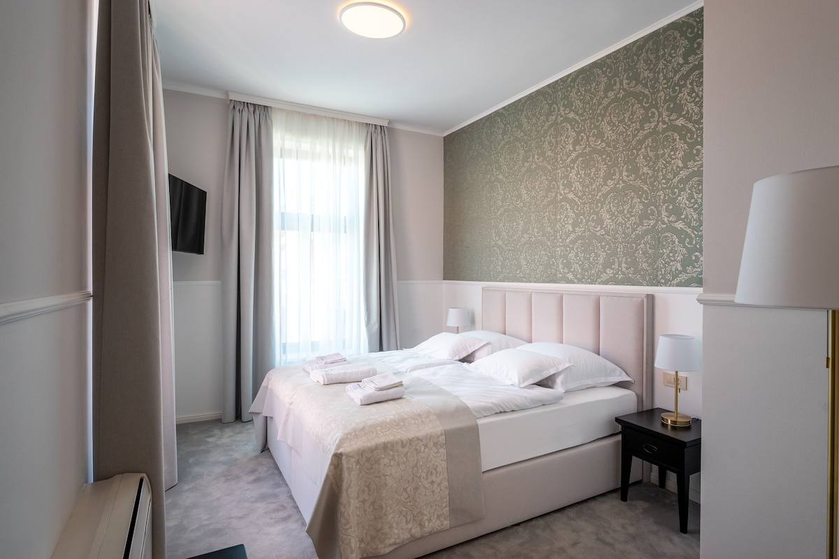 The guest room features a spacious bed adorned with light-colored linens, complemented by fluffy pillows. A subtle patterned wallpaper enhances the ambiance, while natural light filters through sheer curtains. A small nightstand holds a lamp, and a flat-screen television is mounted on the wall.