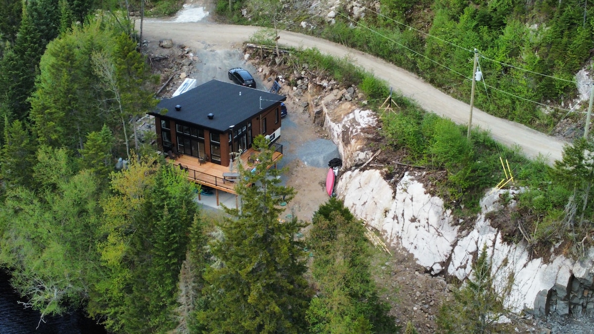 An aerial view of a contemporary chalet is shown, situated amidst greenery. The building features large windows and a spacious terrace. A winding dirt road leads to the property, with a vehicle parked nearby, surrounded by trees and natural landscape.