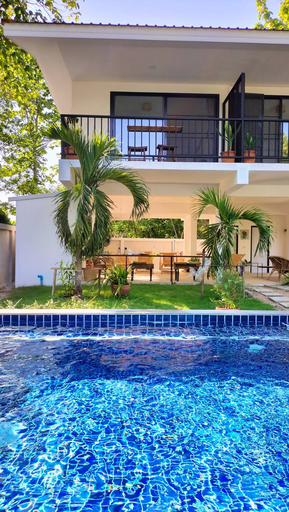 A tranquil outdoor area is highlighted by a sparkling blue pool, with a two-story building visible in the background. Green palm trees frame the scene, and a spacious patio equipped with wooden furniture is situated beneath the balcony.