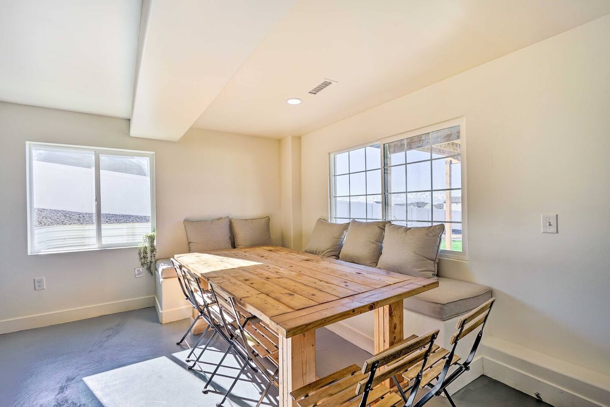 A dining area features a large wooden table surrounded by metal chairs, offering ample seating. Natural light floods in through two windows, creating a bright and open atmosphere. Cozy bench seating is integrated into the corner, enhancing the inviting space.