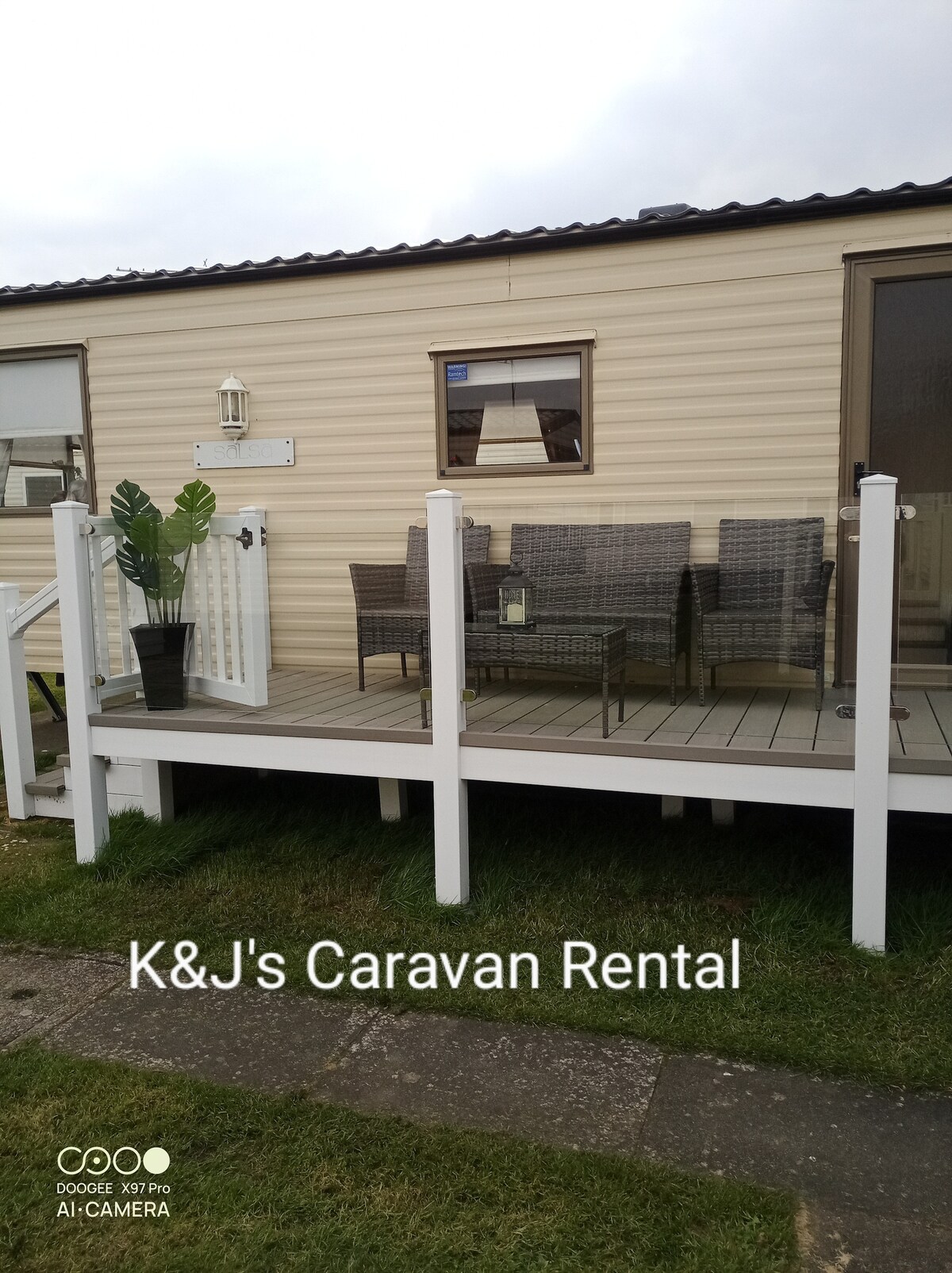 An outdoor seating area is presented on a decking platform, featuring a set of wicker chairs and a table. A potted plant adds a touch of greenery, while the caravan's exterior is shown in neutral tones, complemented by large windows for natural light.