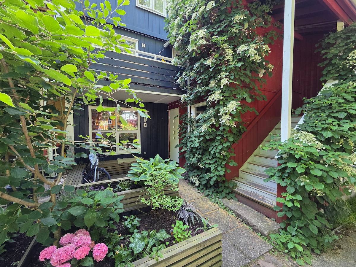 A pathway lined with lush green plants and colorful flowers leads to the entrance of the building. The exterior is framed by vibrant foliage, showcasing a blend of dark and light colors. Stairs are visible, providing access to the upper levels.