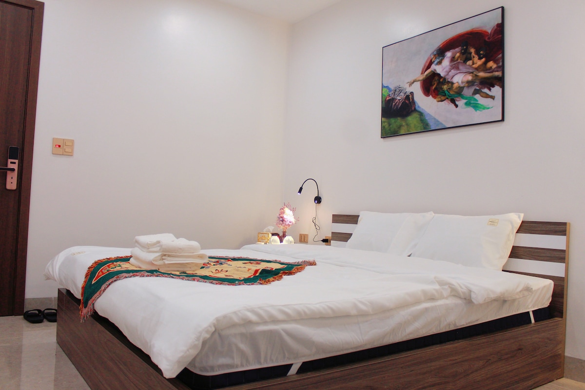 The sleeping area features a spacious bed dressed in crisp white linens. A colorful artwork adorns the wall above. A bedside lamp provides soft illumination, while towels are neatly arranged on the bed, enhancing the room's clean and inviting appearance.