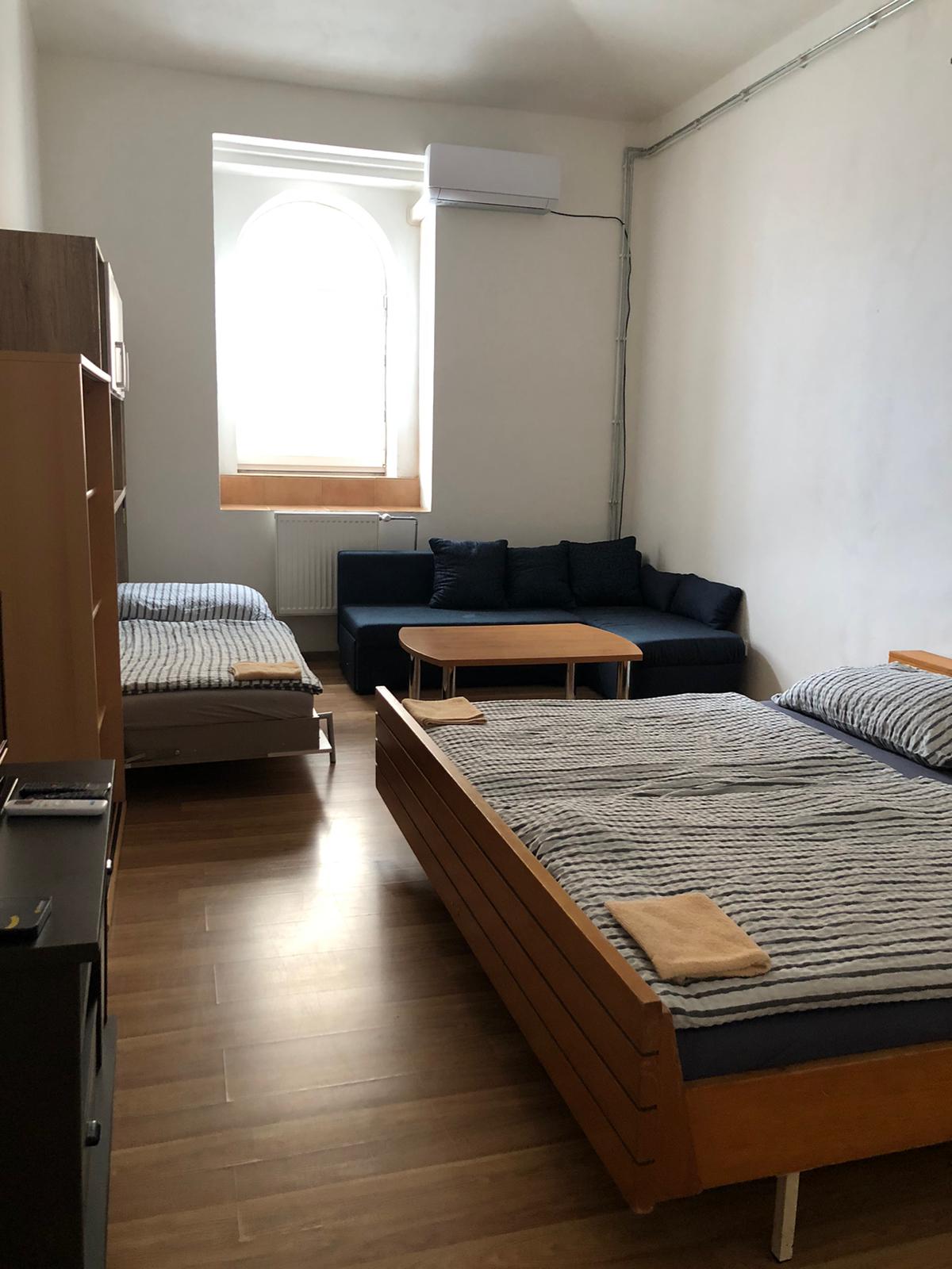 A bright room features two spacious beds with striped bedding positioned alongside a dark sofa and a small table. Natural light enters through an arched window, illuminating the wooden floor and simple wooden furniture.