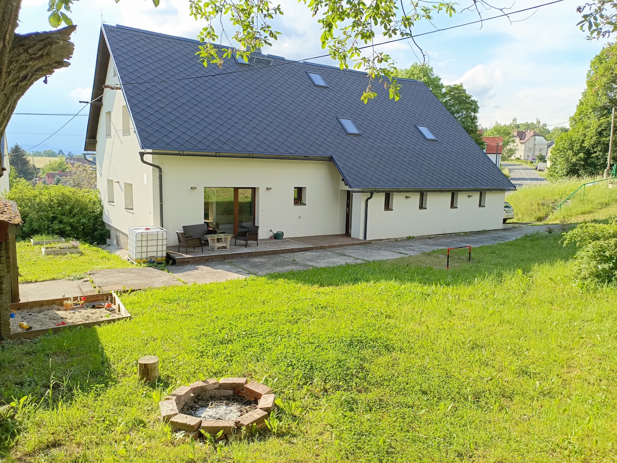 A spacious exterior view of the villa showcases a well-maintained yard and a patio area furnished with seating. The structure features a sloped roof with skylights and large windows that invite natural light. A fire pit surrounded by brick is visible in the grass.