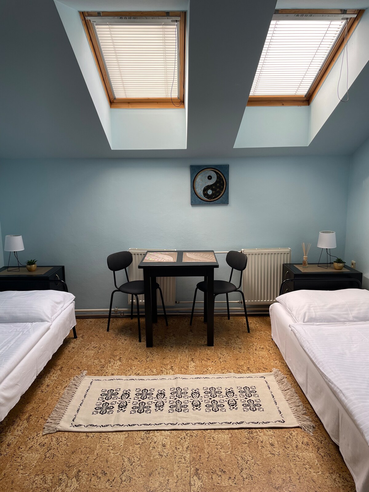 A well-lit room features two single beds adorned with white linens, positioned beside nightstands with small lamps. A small dining table is situated in the center, accompanied by two chairs. Natural light filters in through skylights above, creating a bright ambiance.