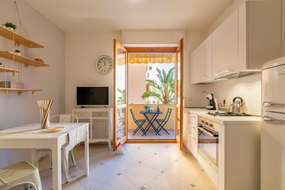 A bright kitchen area features a white dining table and chairs, complemented by open shelving. A vintage clock is mounted on the wall, while a large window with opened doors leads to a small balcony with a table and chairs, surrounded by green plants.