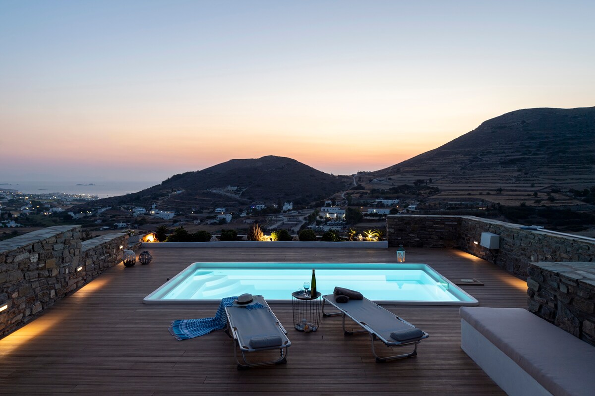 A private swimming pool is surrounded by a wooden deck, featuring two lounge chairs and a small table. The view overlooks hills and the distant sea at sunset, with soft hues in the sky creating a serene atmosphere.