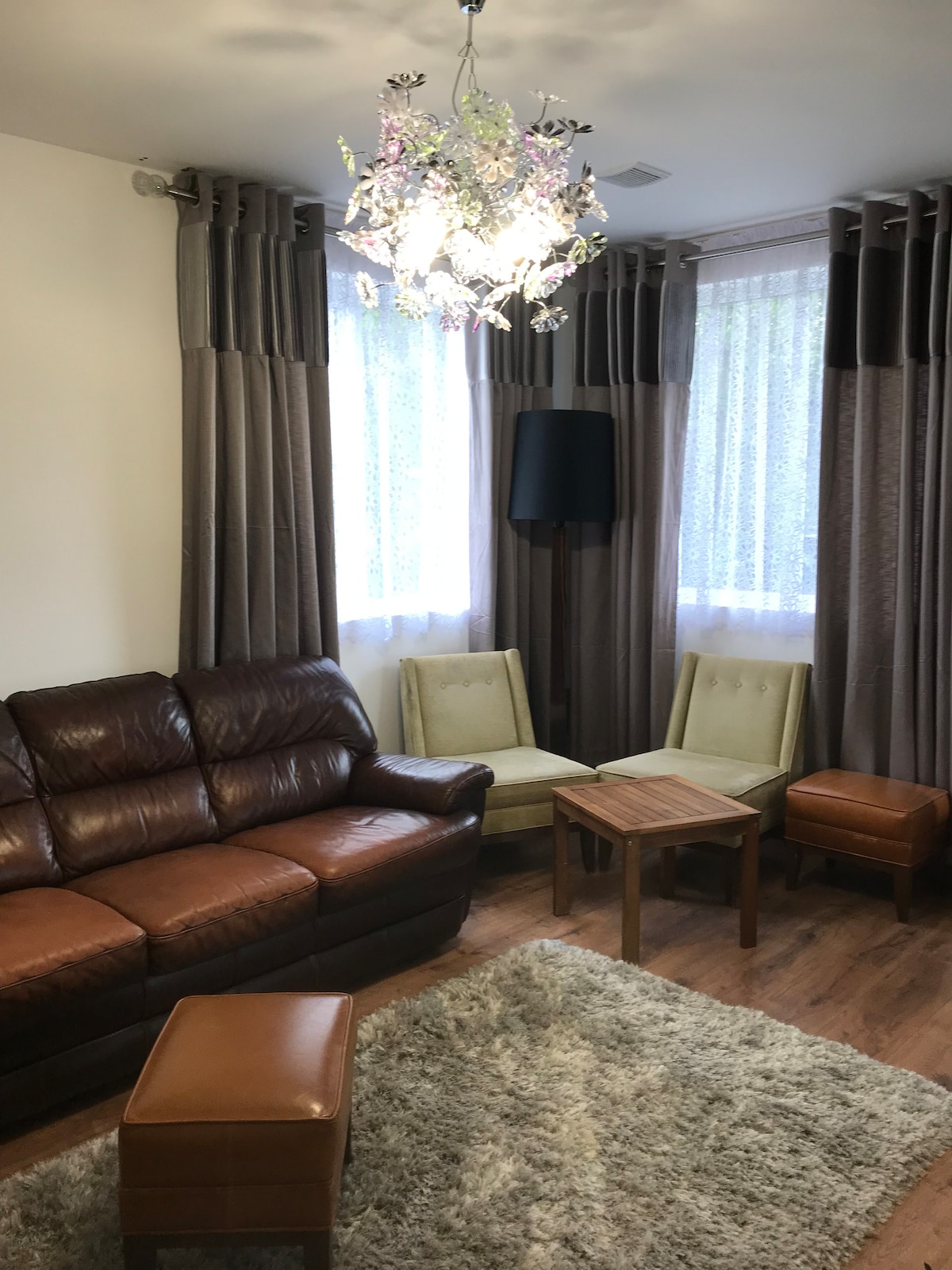 A living area features a brown leather sofa paired with two light green armchairs, positioned next to a wooden table. A plush rug covers the floor, while sheer curtains allow natural light to filter in, creating a comfortable and bright space.