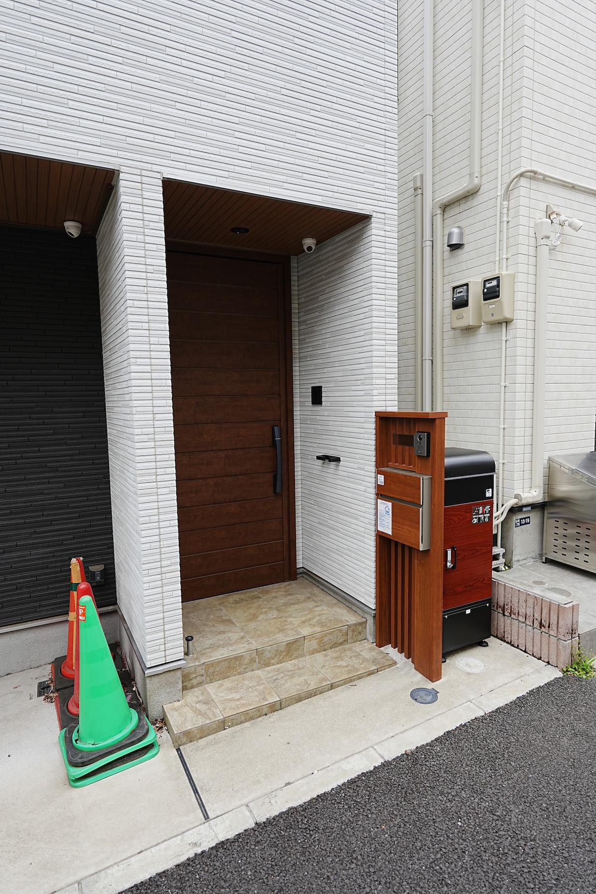 The entrance showcases a modern wooden door framed by textured white walls. A small set of stone steps leads to the door, highlighted by a mail slot and a nearby mailbox. Green traffic cones are positioned on the side, contributing to the minimalistic exterior.